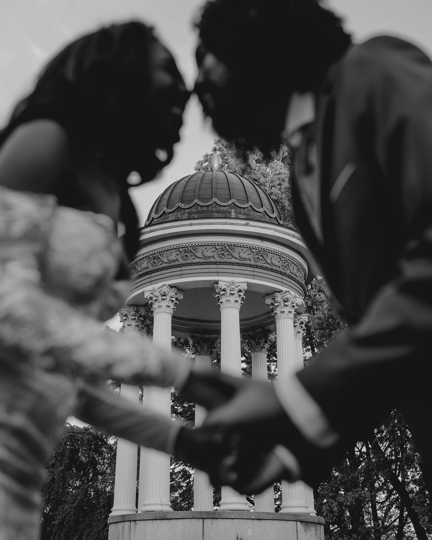 Black and white photo of bride and groom holding hands with ornate domed gazebo in background.