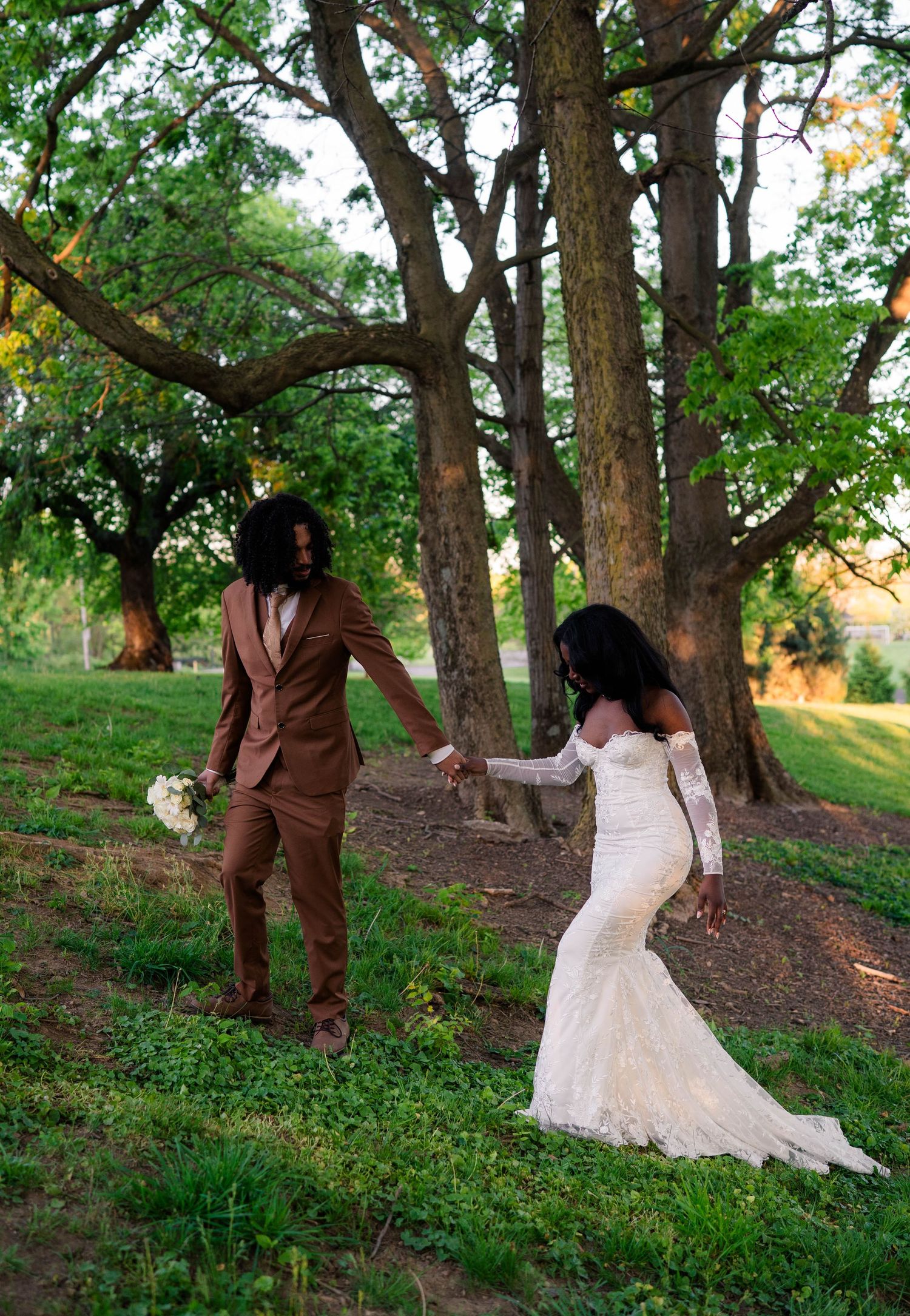 A Black bride and groom hold hands walking through a lush green park, she in lace gown, he in brown suit.