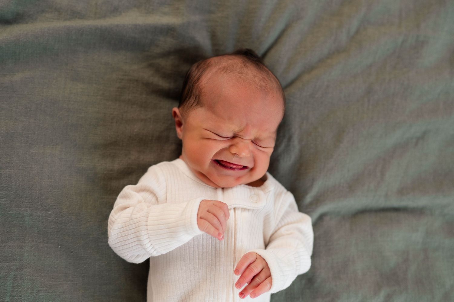 Close-up of a crying newborn baby in white onesie lying on a gray blanket.