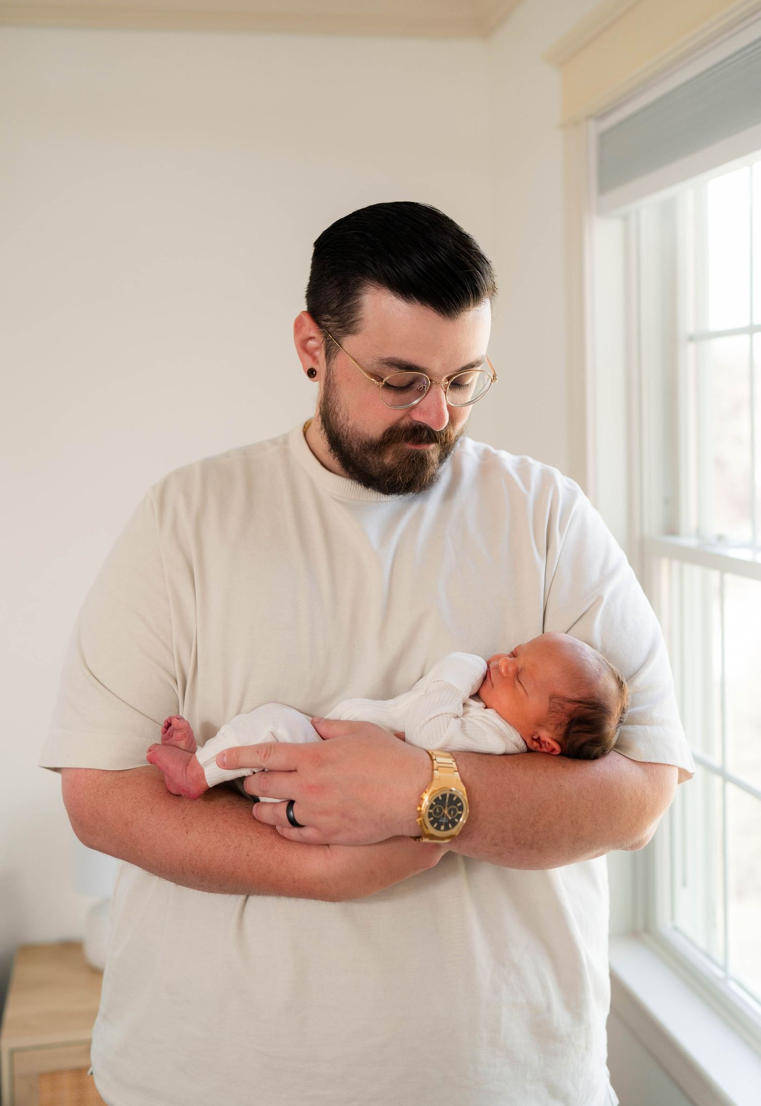 Bearded father in white t-shirt gently holds sleeping newborn baby near a bright window at home.