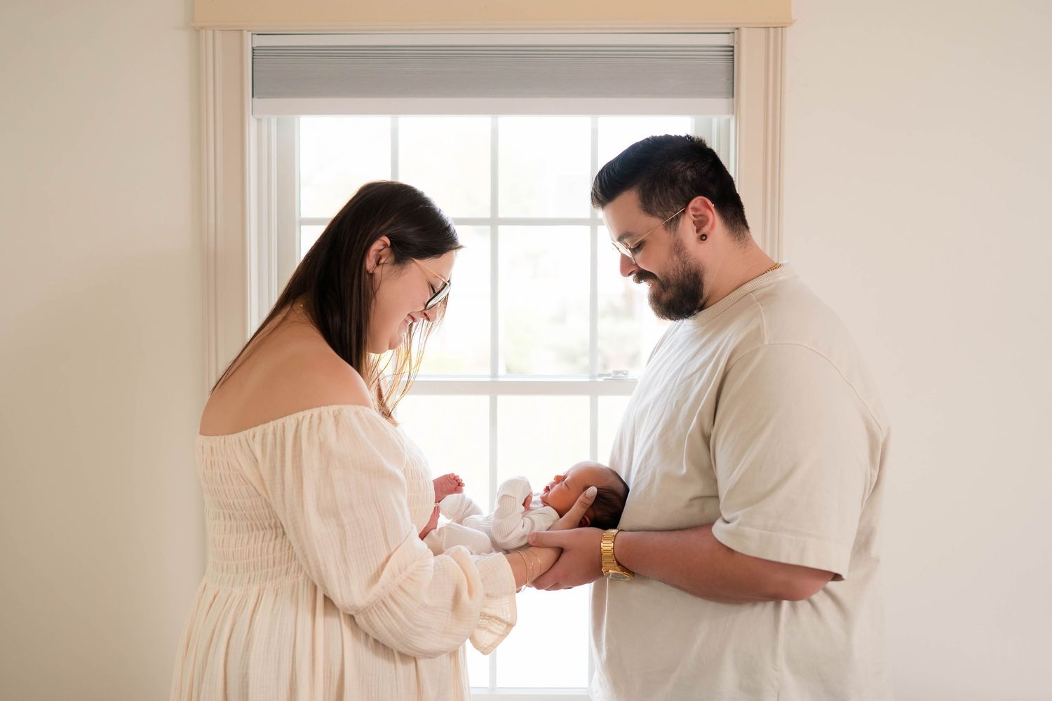 New parents lovingly hold their newborn baby together by a bright window in a tender family portrait.