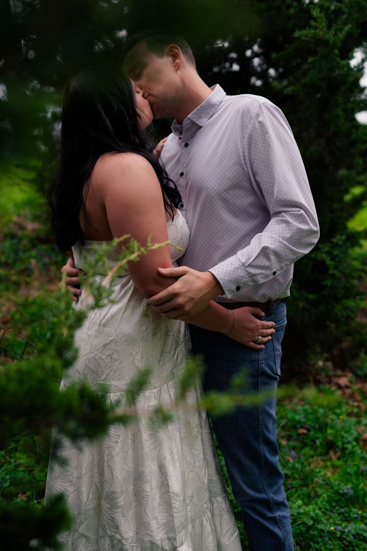 Couple sharing a romantic kiss outdoors surrounded by lush green foliage, woman in white dress, man in blue shirt.