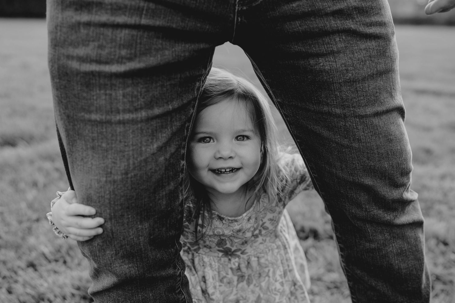 Black and white photo of a smiling toddler girl peeking through an adult's legs outdoors.