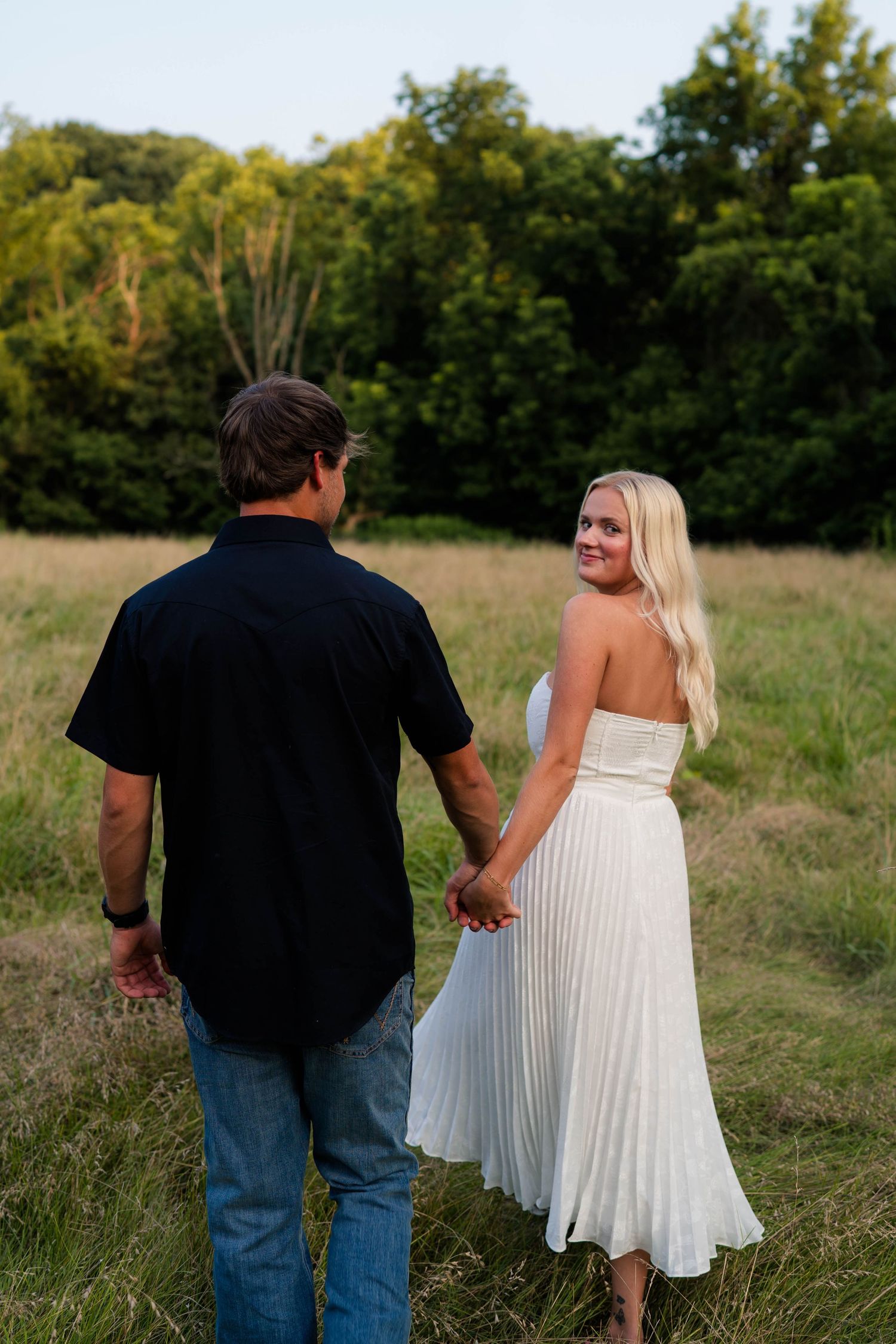 Couple holding hands in a sunlit meadow, blonde woman in white dress glancing back, lush green trees behind.