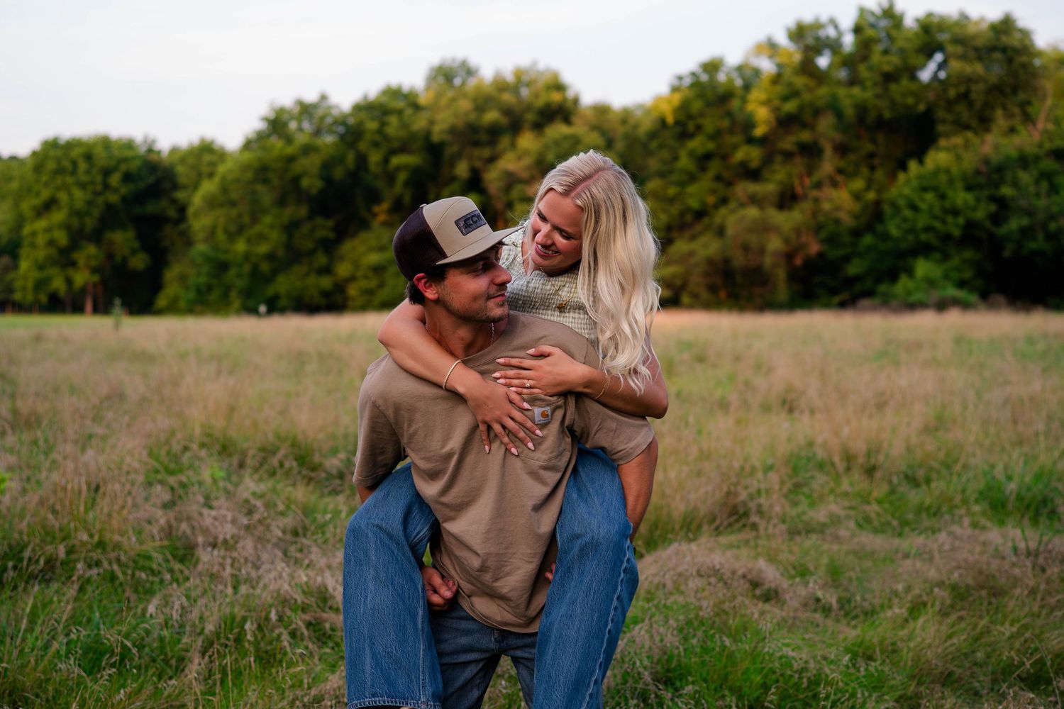 A couple laughs joyfully in a grassy field, man giving woman a piggyback ride, both wearing casual clothes.