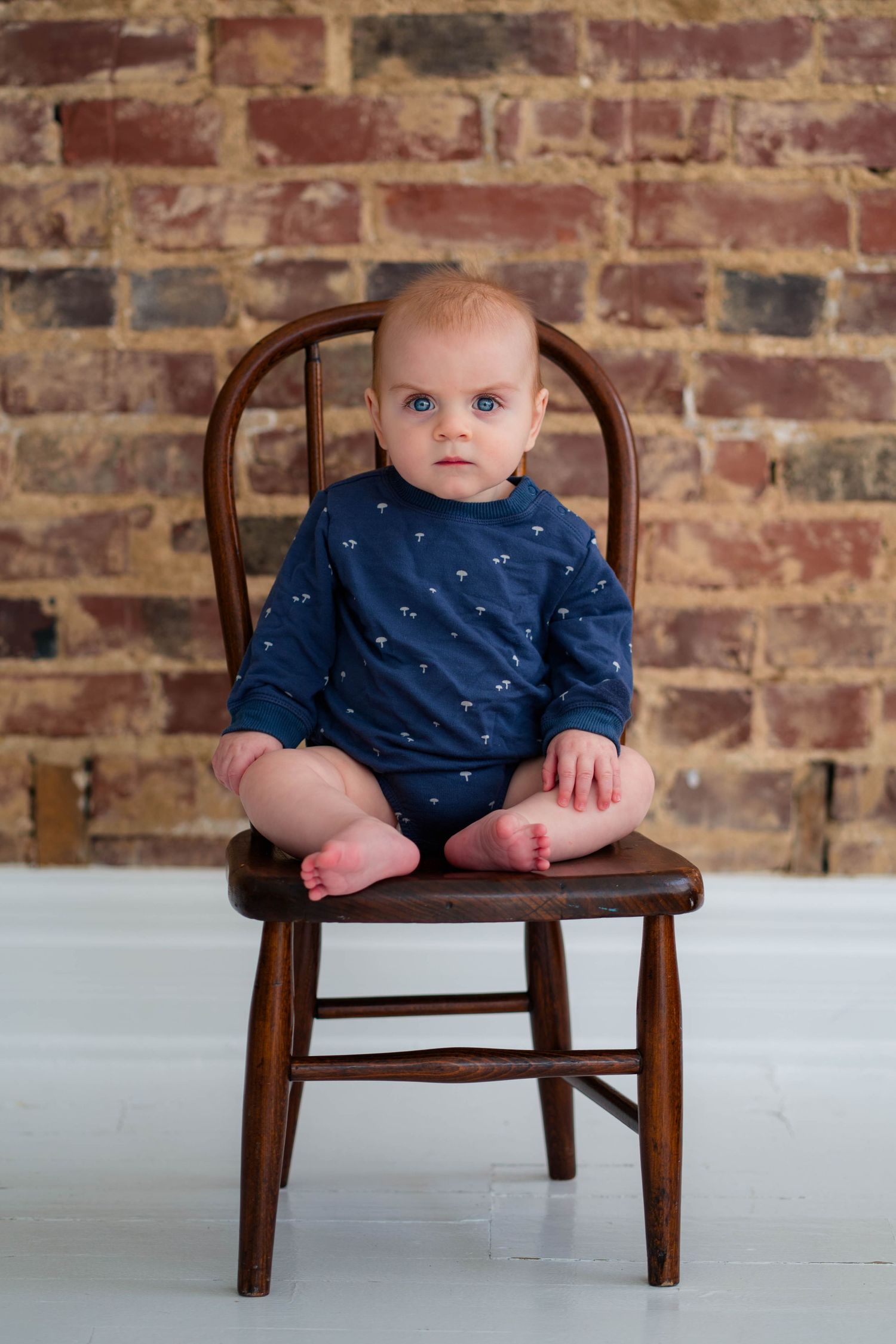 Blue-eyed baby in navy patterned onesie sitting on wooden chair against rustic brick wall.