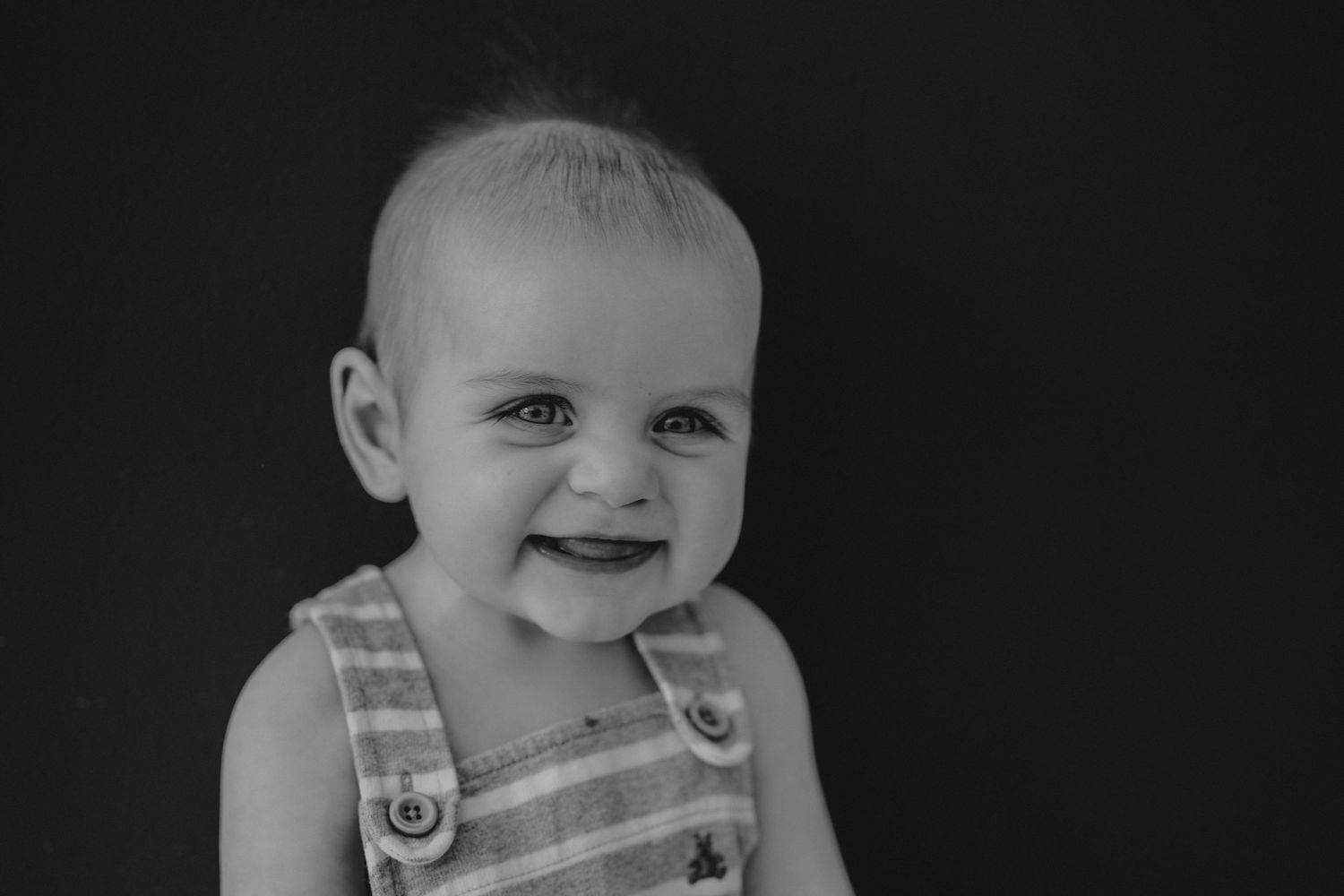 Black and white close-up portrait of smiling baby in striped overalls against dark background.