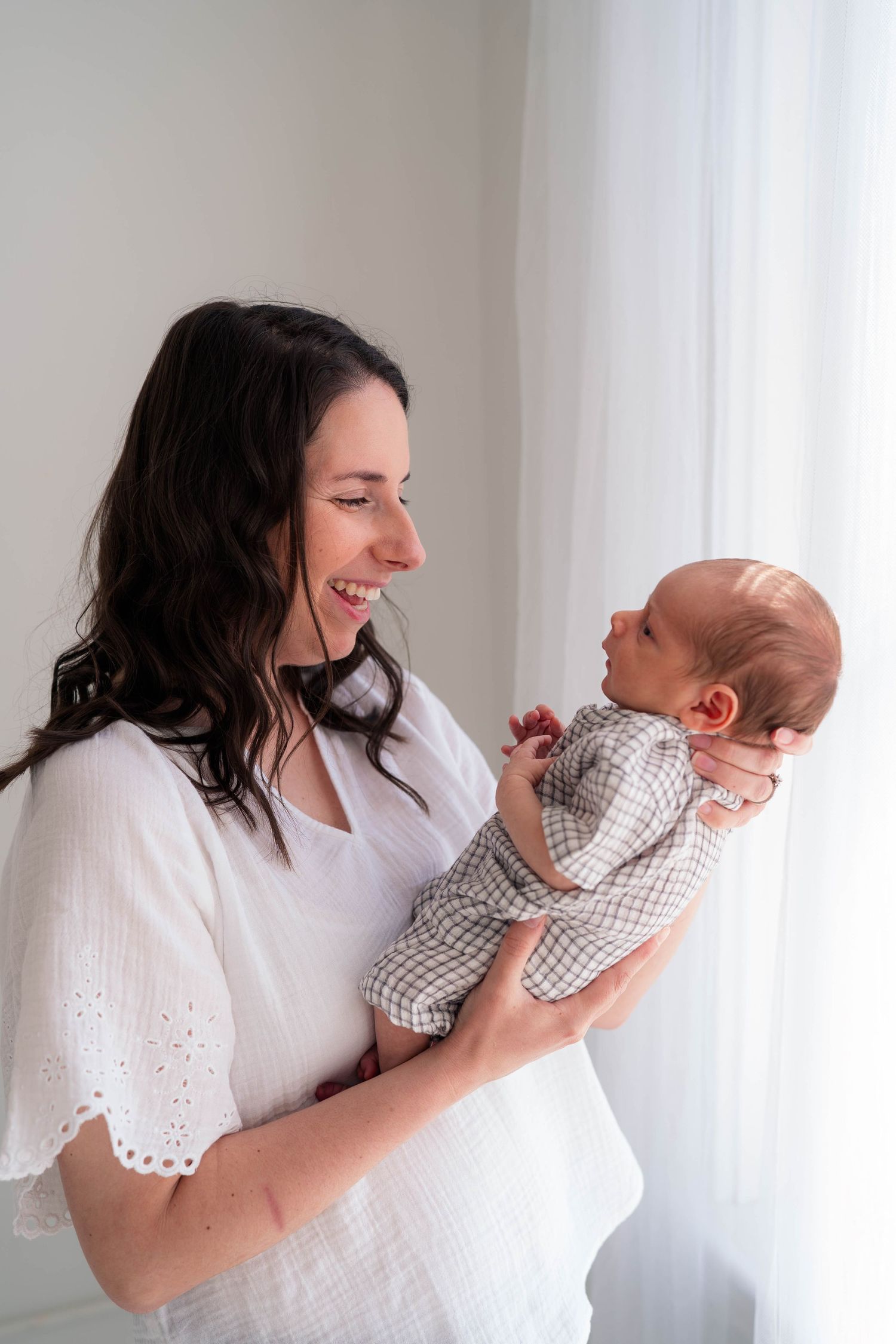 Smiling dark-haired mother in white dress holding her newborn baby near a bright window.