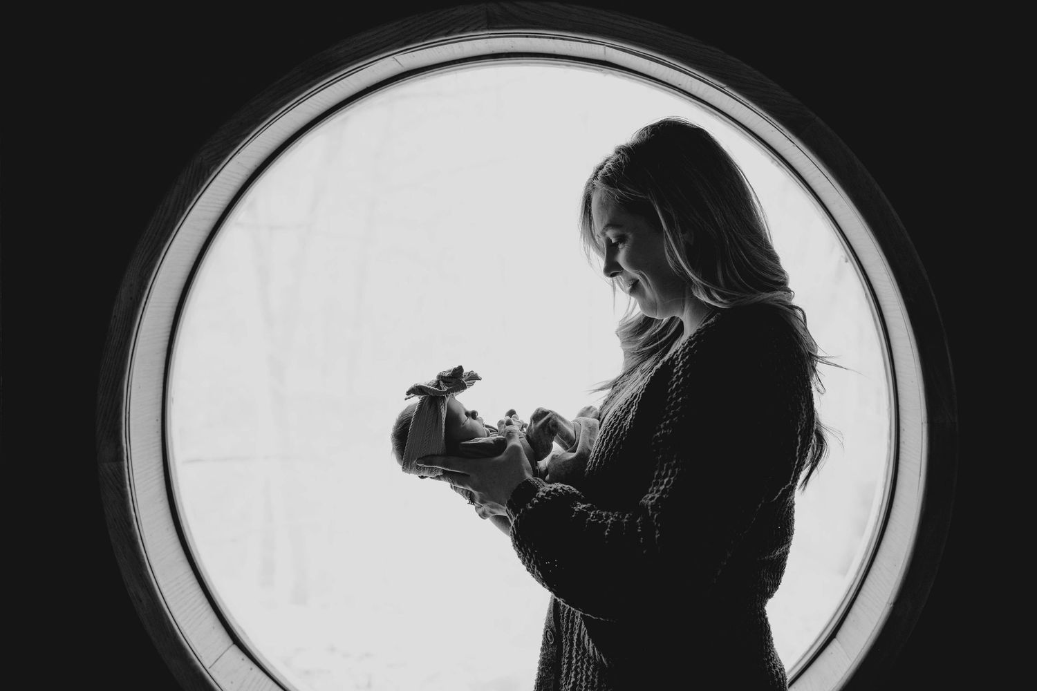 Black and white silhouette of a mother gazing lovingly at her newborn in front of a round window.