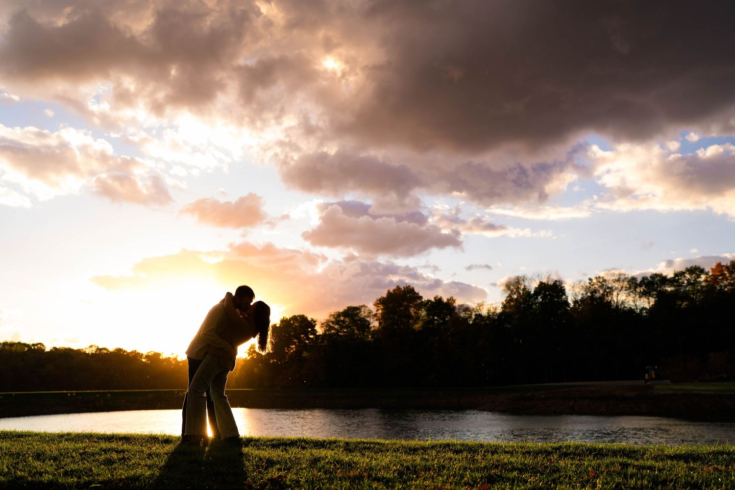 Romantic couple silhouette kissing at sunset by a lake with dramatic cloudy sky.