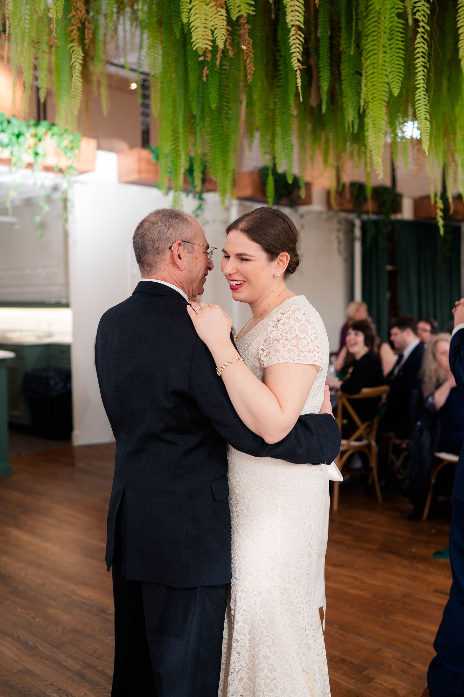 Smiling bride in lace gown shares emotional father-daughter dance at wedding reception.
