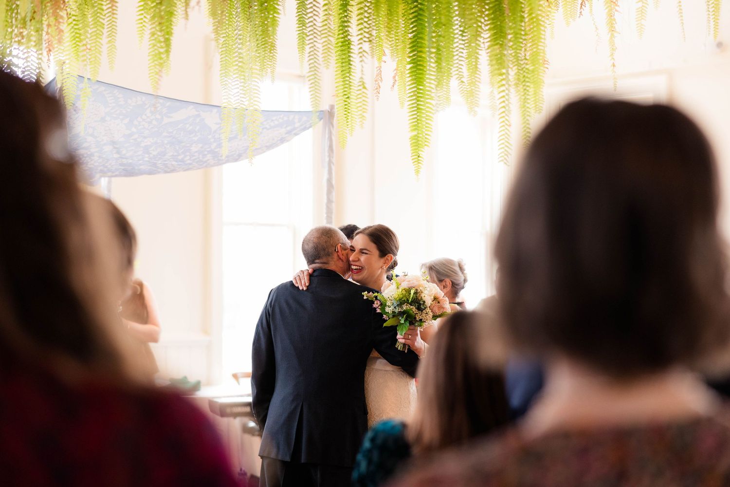 Bride and groom share first kiss at indoor wedding ceremony beneath hanging fern decorations.