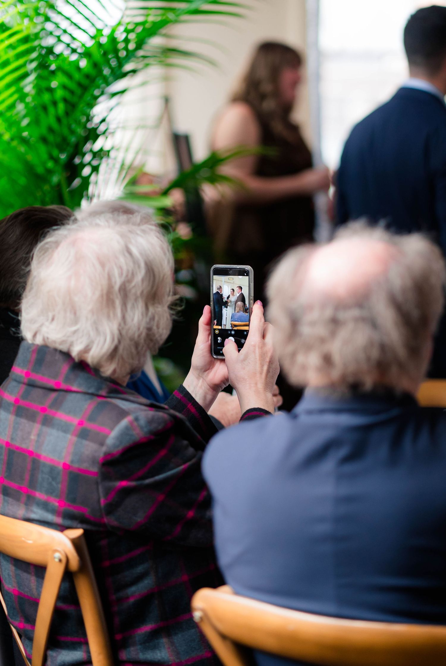 Elderly wedding guest photographs the ceremony on a smartphone while seated indoors.