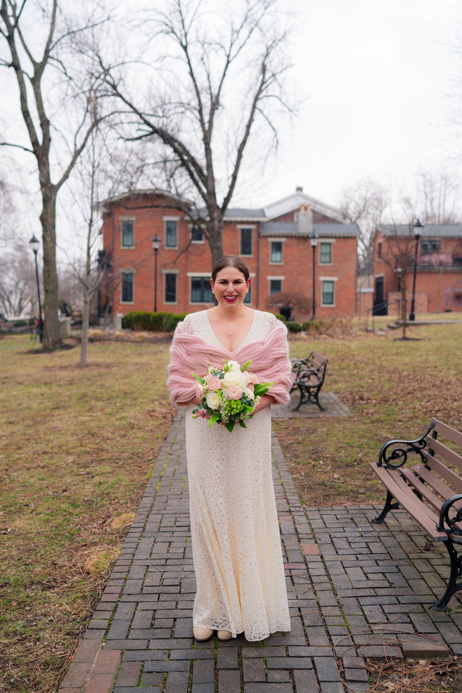 Bride in white lace gown and pink shawl holding a bouquet on a brick path in front of a red brick building.