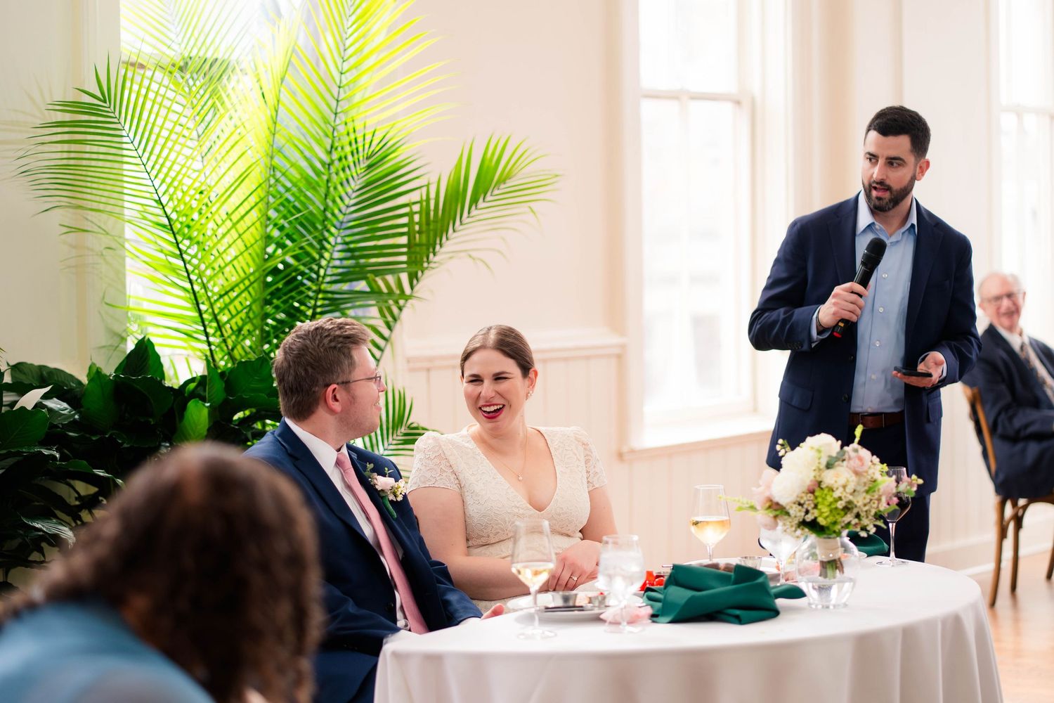 Best man gives toast as bride and groom smile at intimate wedding reception table.