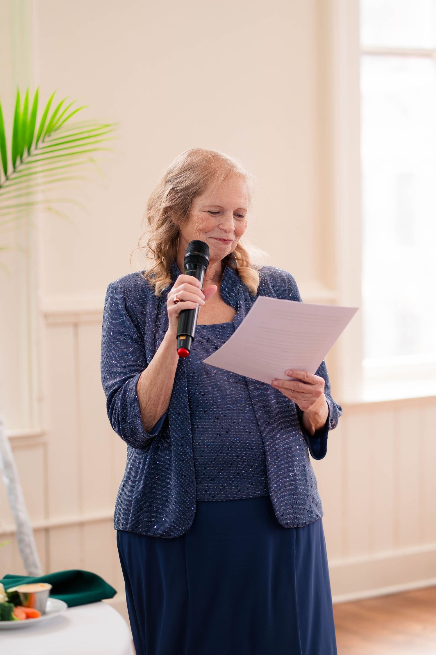Woman in blue dress reads from paper while speaking into microphone at wedding reception.