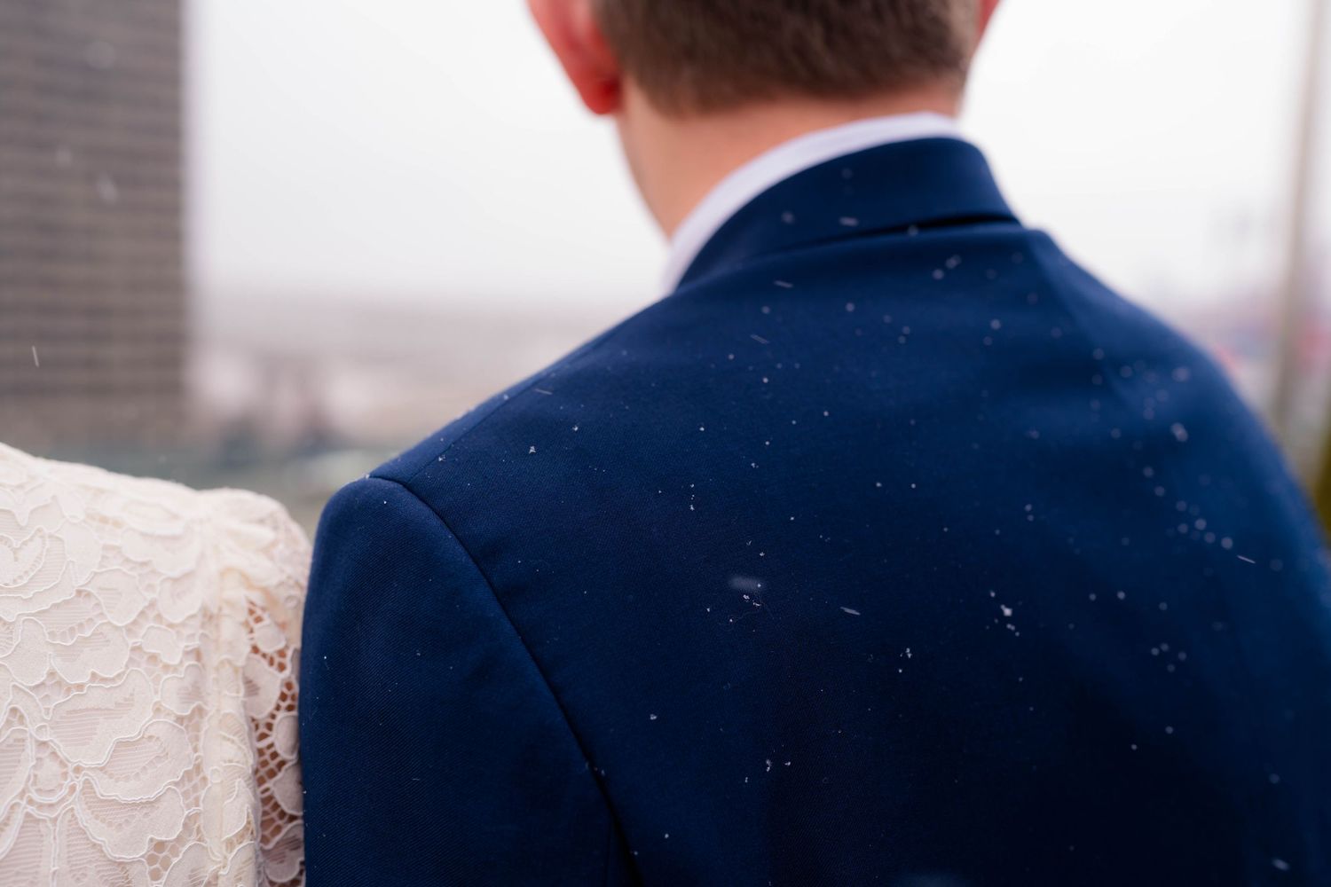Close-up of groom in navy blue suit with snow falling on his back beside bride in white lace dress.