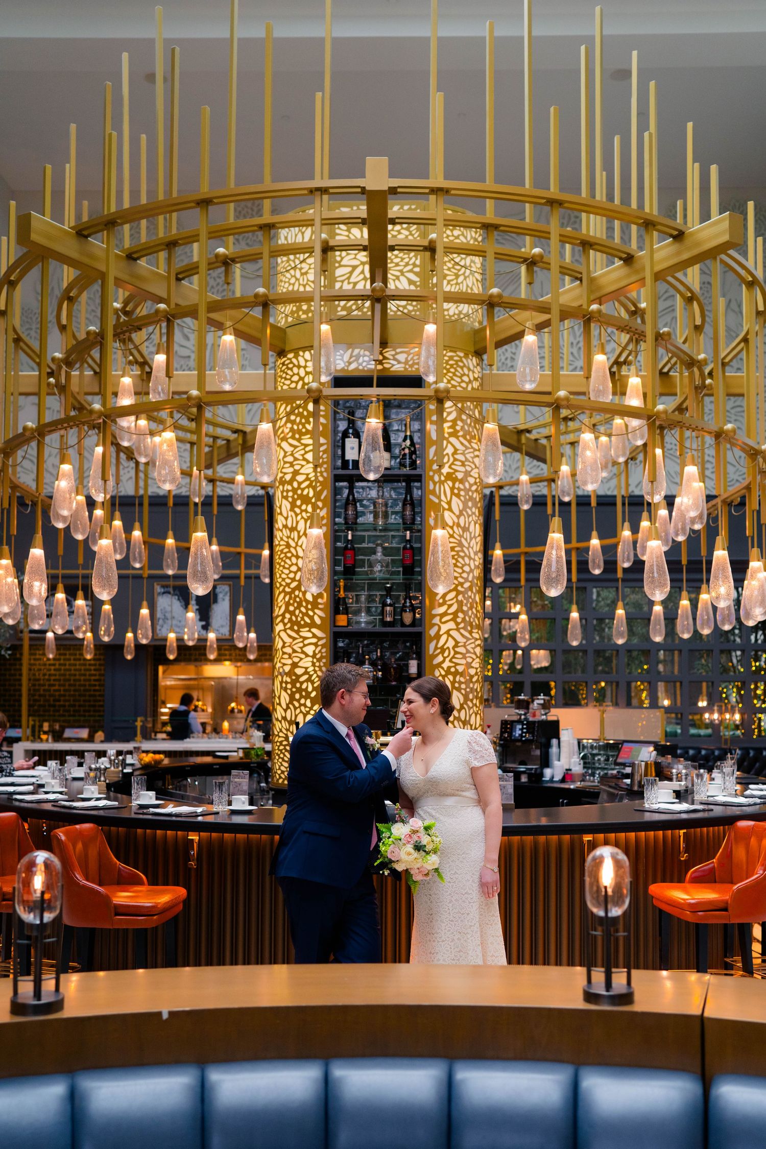 Bride and groom standing beneath an elegant gold chandelier in a luxurious restaurant bar wedding venue.