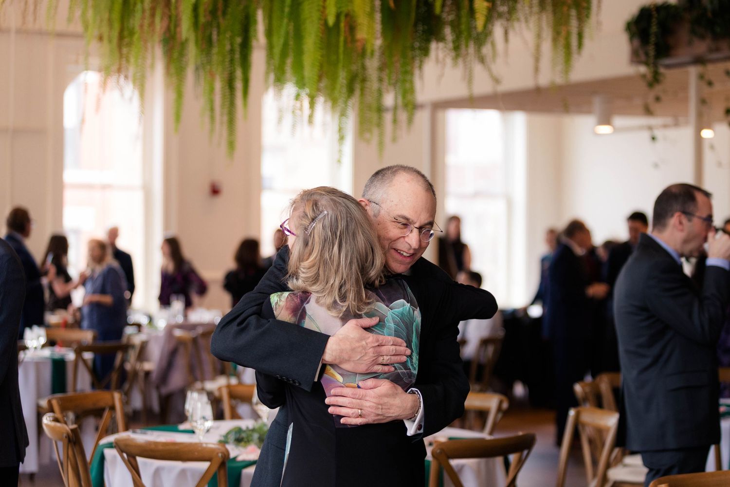 Smiling man embracing a guest at a wedding reception with hanging greenery decor.
