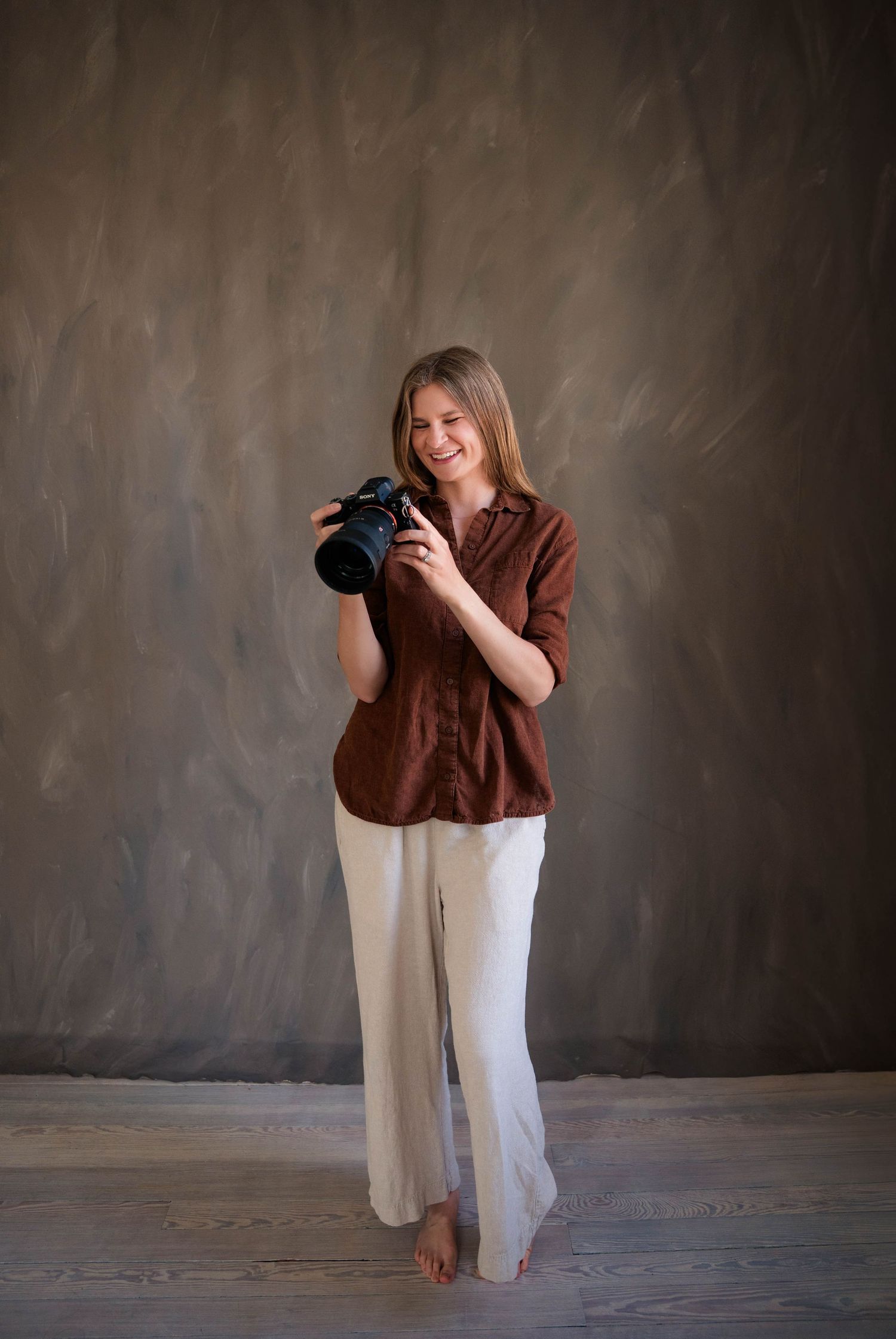 Smiling female photographer standing with camera against textured gray studio backdrop.