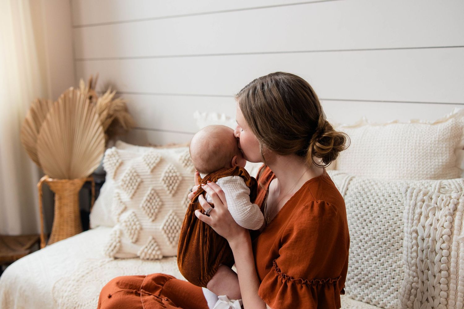 Mother in rust orange dress kissing her newborn baby on a cream boho-styled couch.