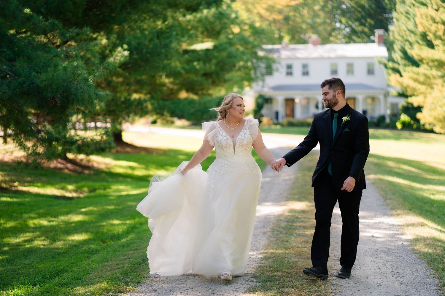 Bride and groom walk hand-in-hand on a tree-lined path outside a white estate home.