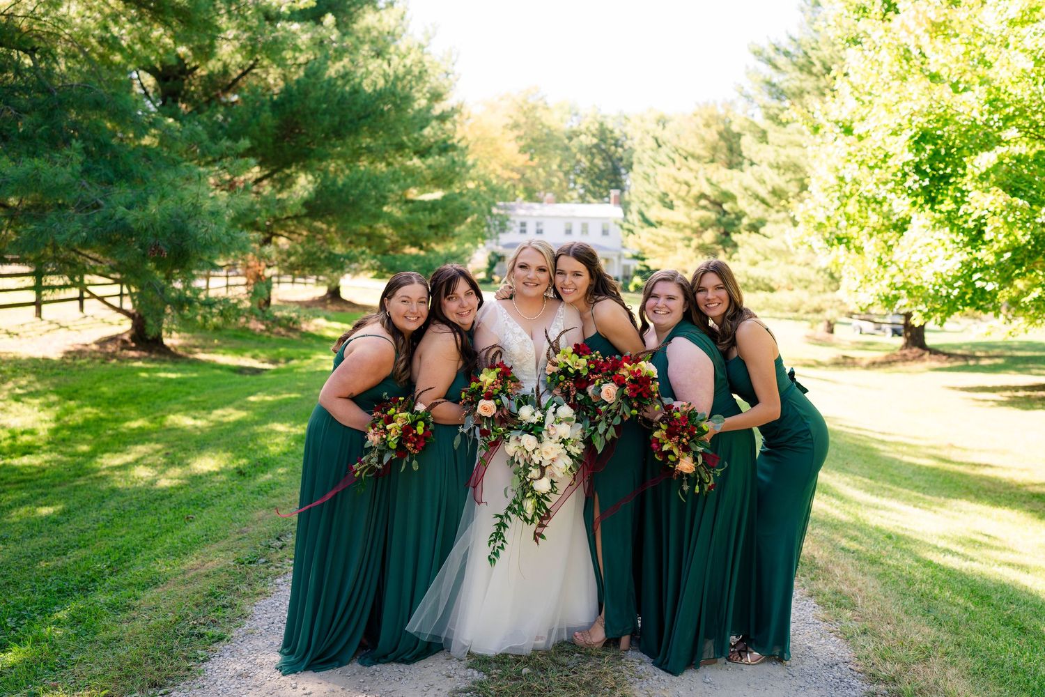 Bride and bridesmaids in emerald green dresses holding vibrant bouquets pose outdoors at a wedding venue.