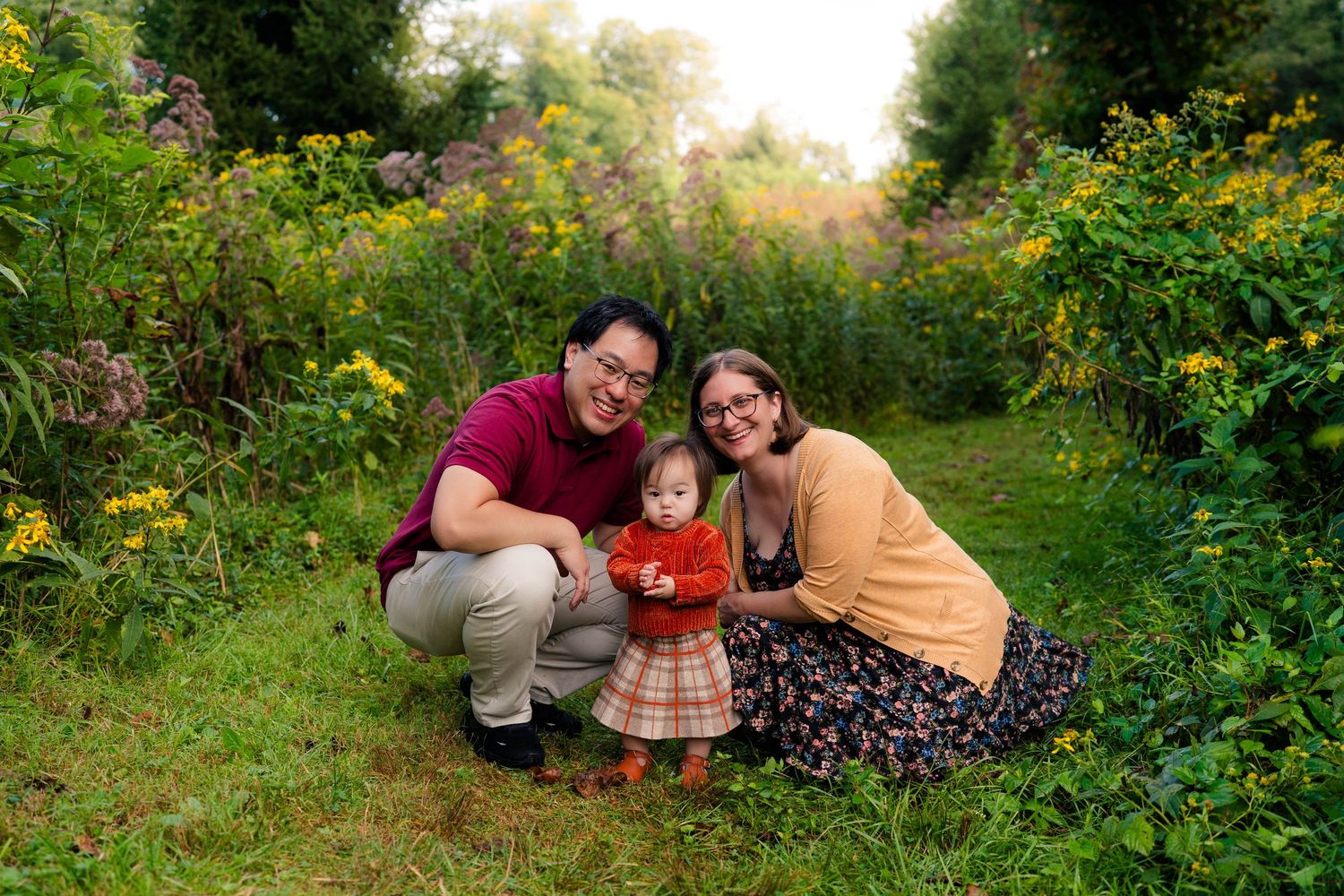 Family of three posing outdoors in a lush garden with yellow wildflowers, parents crouching beside toddler in red outfit.