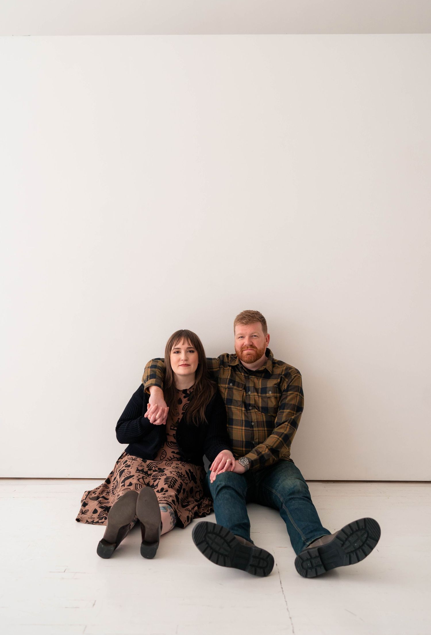 A couple sits casually against a white wall, holding hands in a minimalist indoor space.