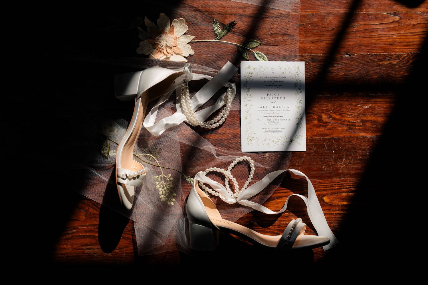 Wedding flat lay featuring bridal heels, pearl jewelry, and invitation on rustic wood surface.