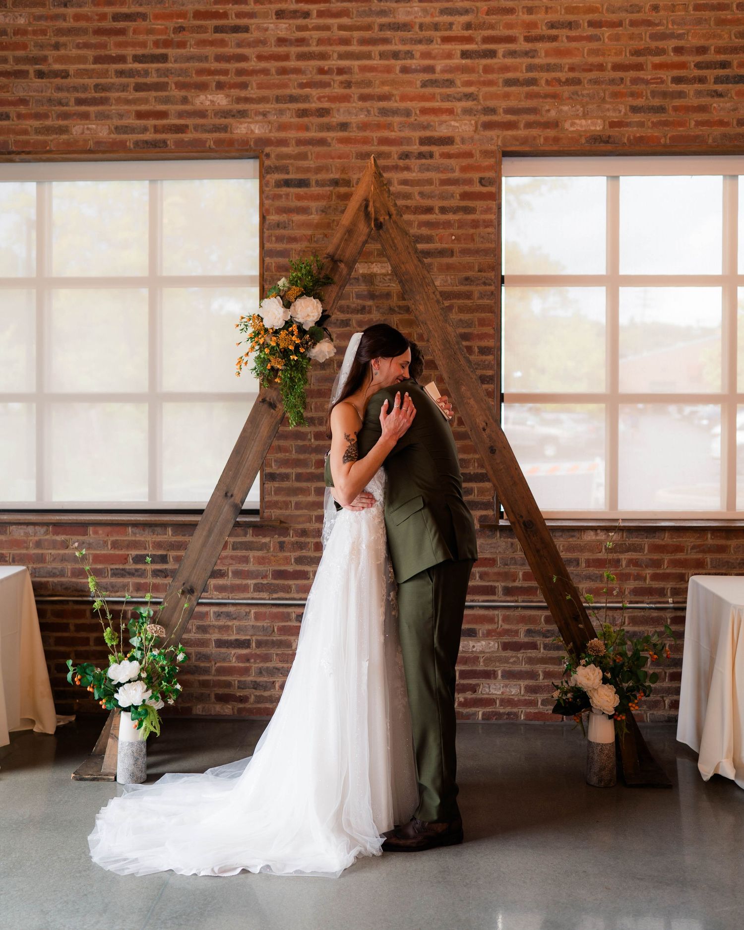 Bride and groom share first kiss under rustic wooden triangular arch with floral accents in brick ceremony venue.