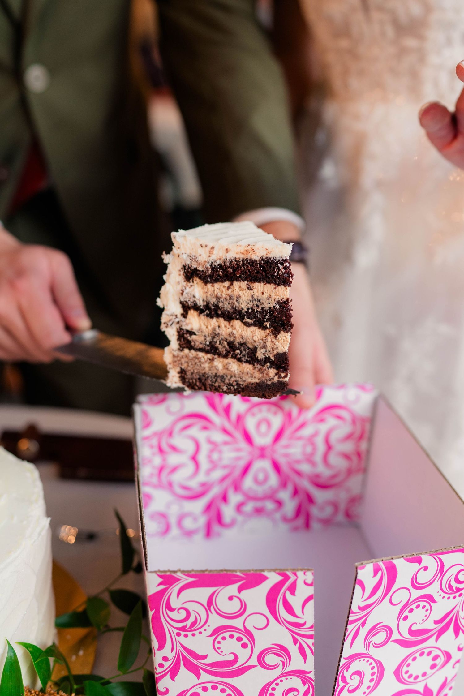 Close-up of a layered chocolate wedding cake slice being served on a decorative pink and white box.