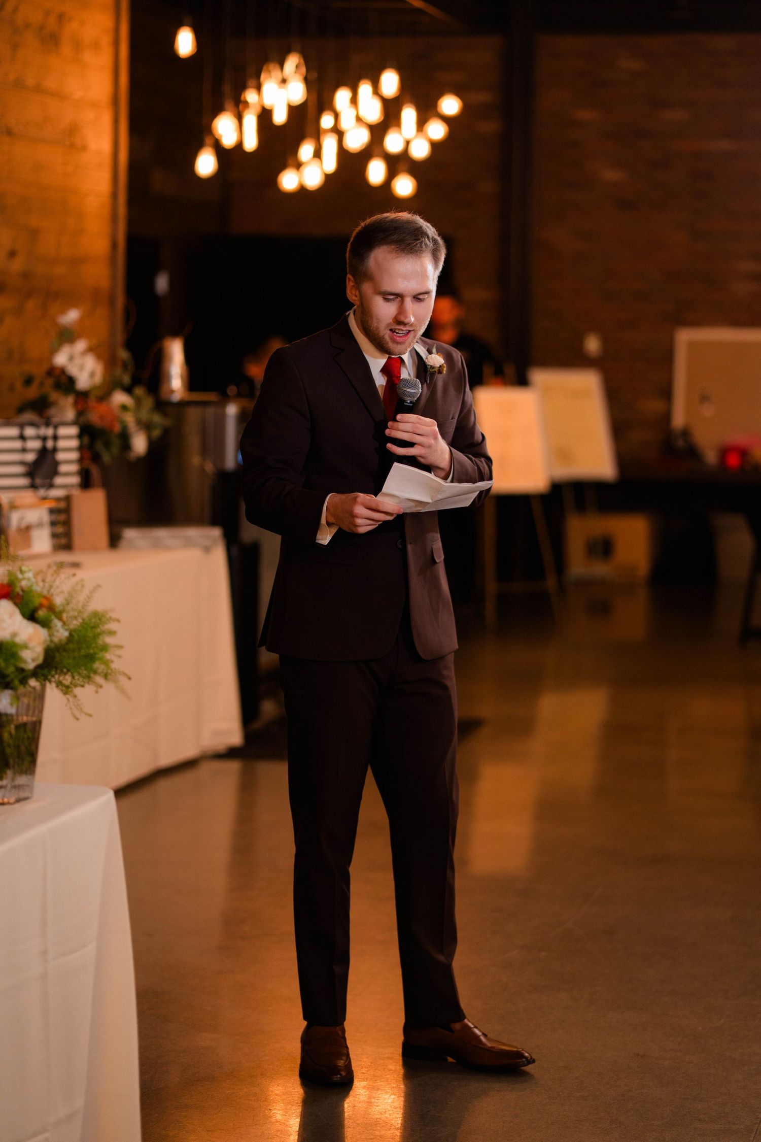 Groom in dark suit reads speech from paper while holding microphone at wedding reception.
