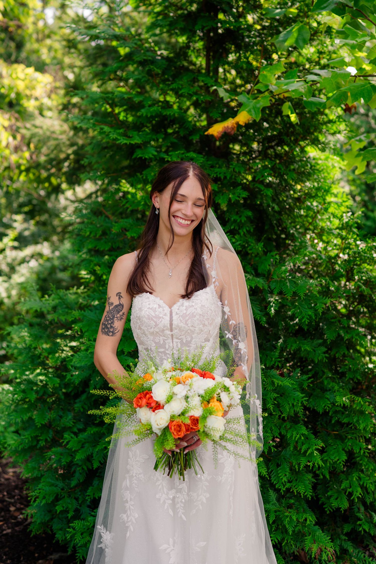 Smiling bride in lace gown holding orange and white bouquet surrounded by green trees.