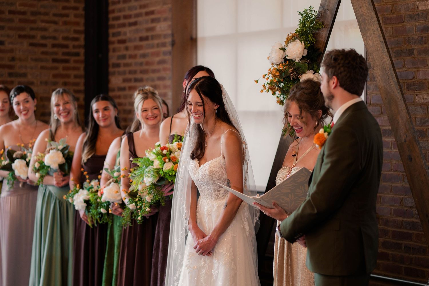 Bride smiling during wedding ceremony with bridesmaids holding floral bouquets in a rustic brick venue.