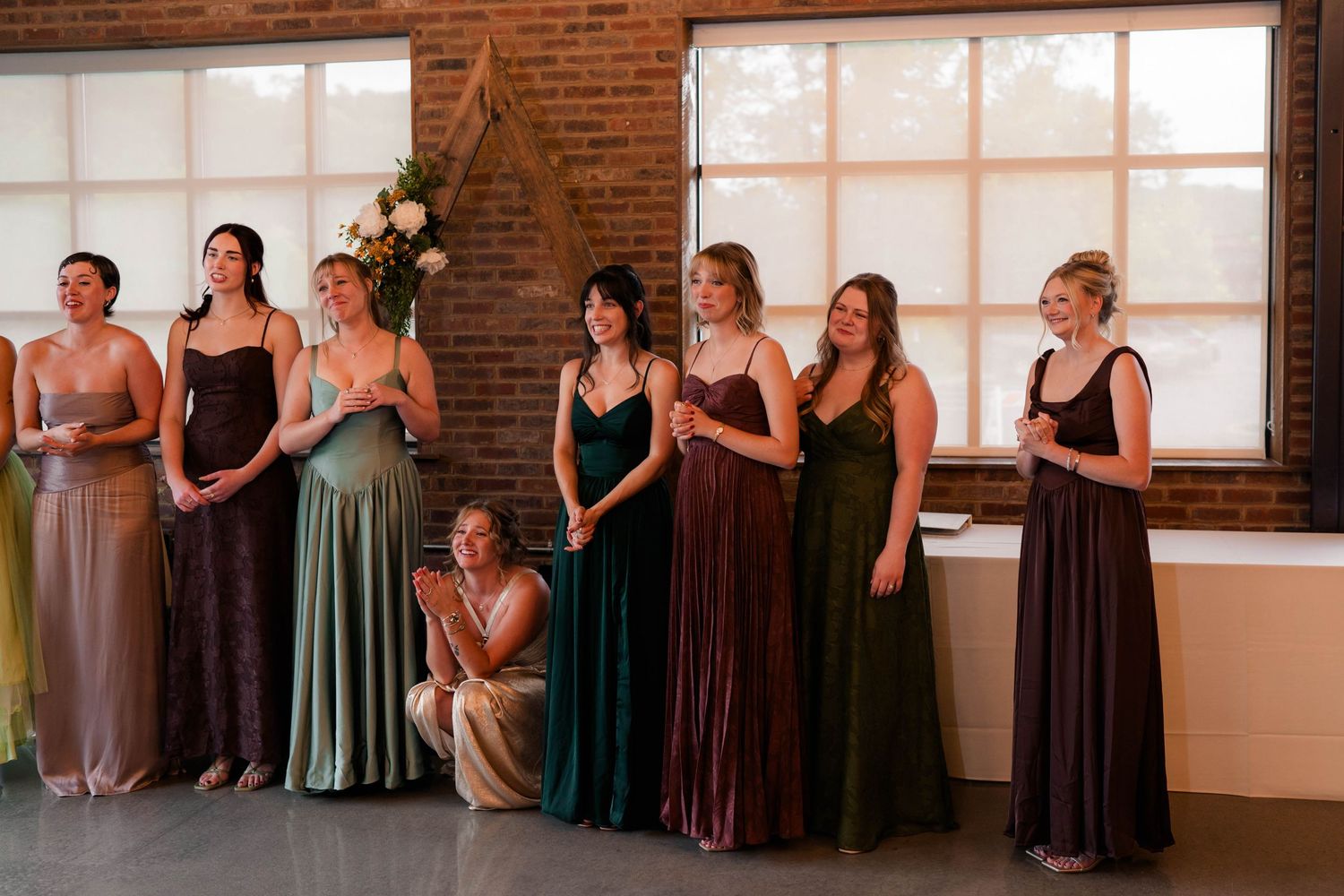 Bridesmaids in mismatched earth-tone velvet gowns standing together in a rustic brick ceremony venue.