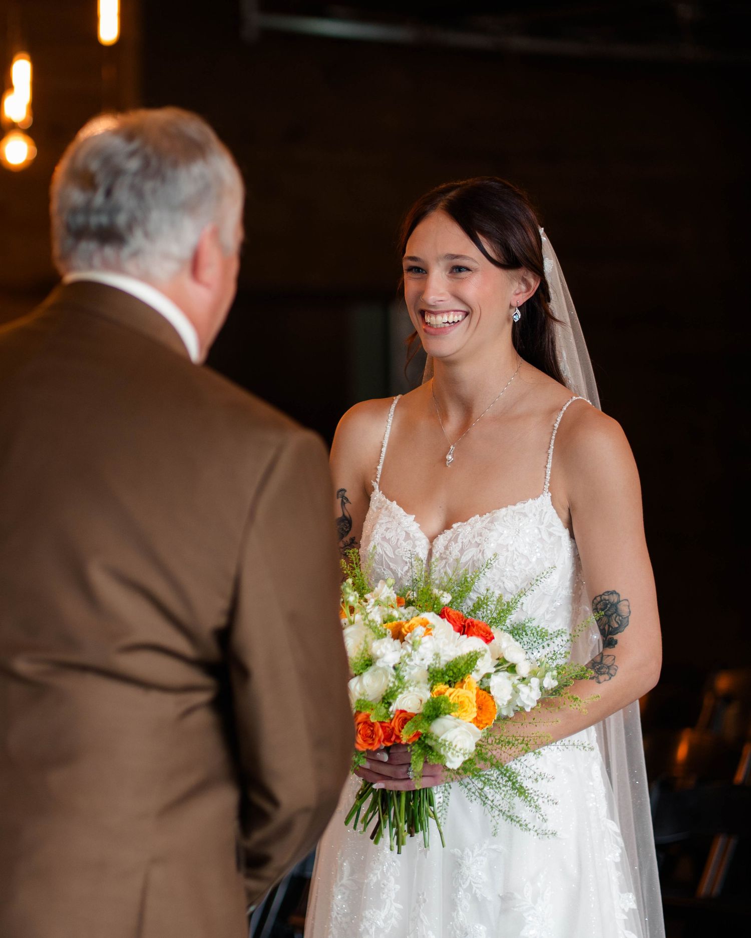 Radiant bride in lace gown holding orange and white bouquet sharing emotional first look moment.