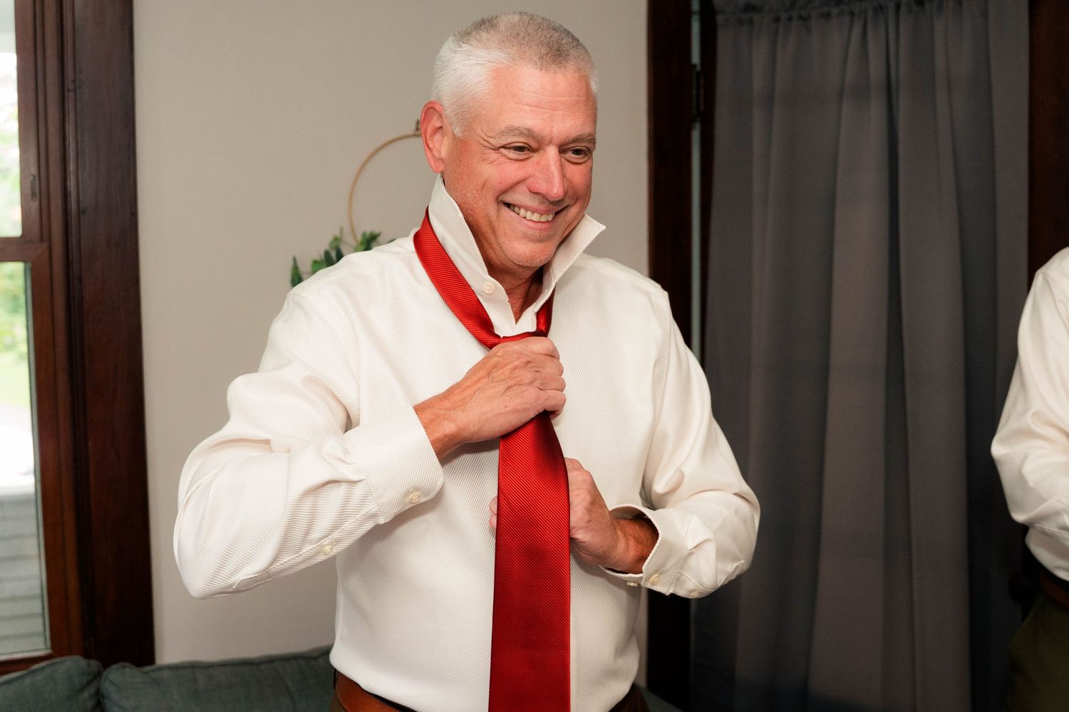 Smiling older man in white dress shirt adjusting red tie, getting ready for formal event.