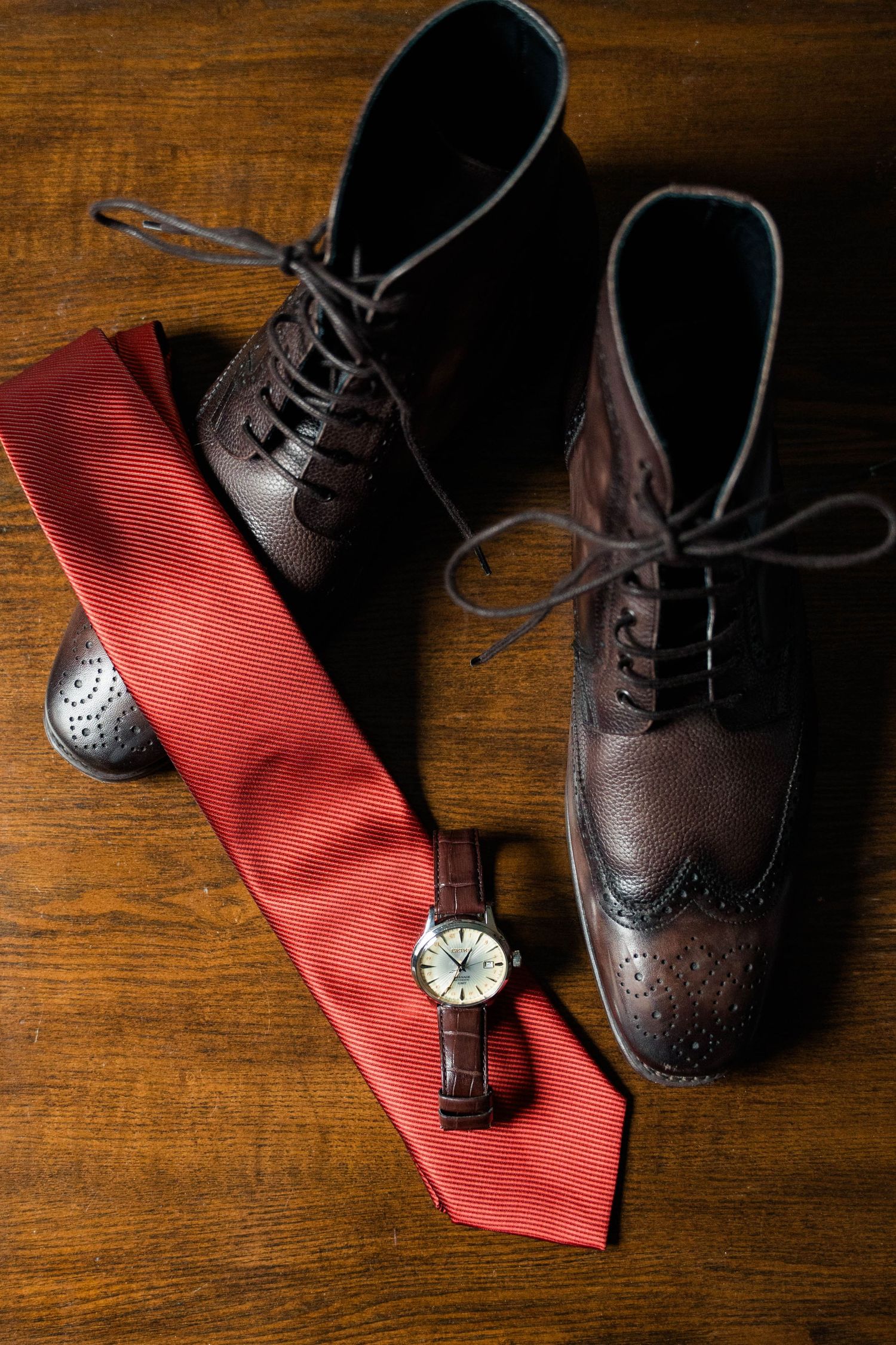 Brown leather brogue dress shoes with red tie and analog watch on wooden floor.