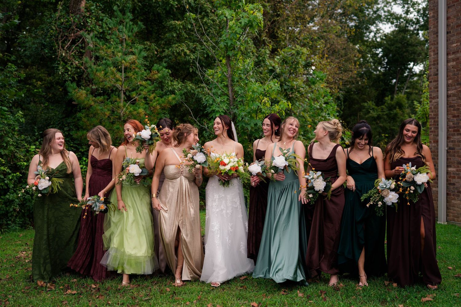 Bride surrounded by bridesmaids in mismatched green and burgundy gowns holding bouquets on a grassy lawn.