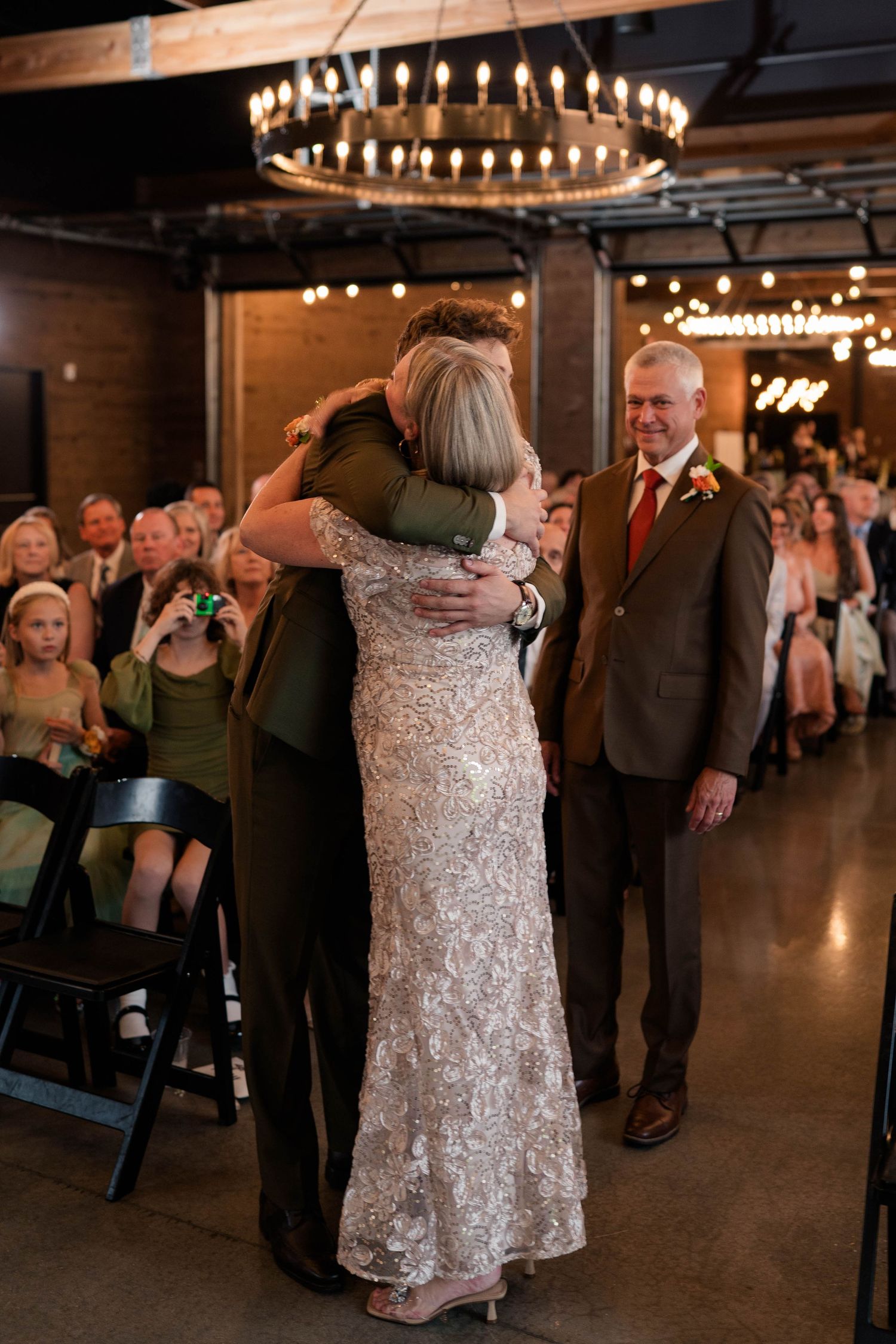 Groom hugging mother in sequin dress during wedding ceremony processional at indoor venue.