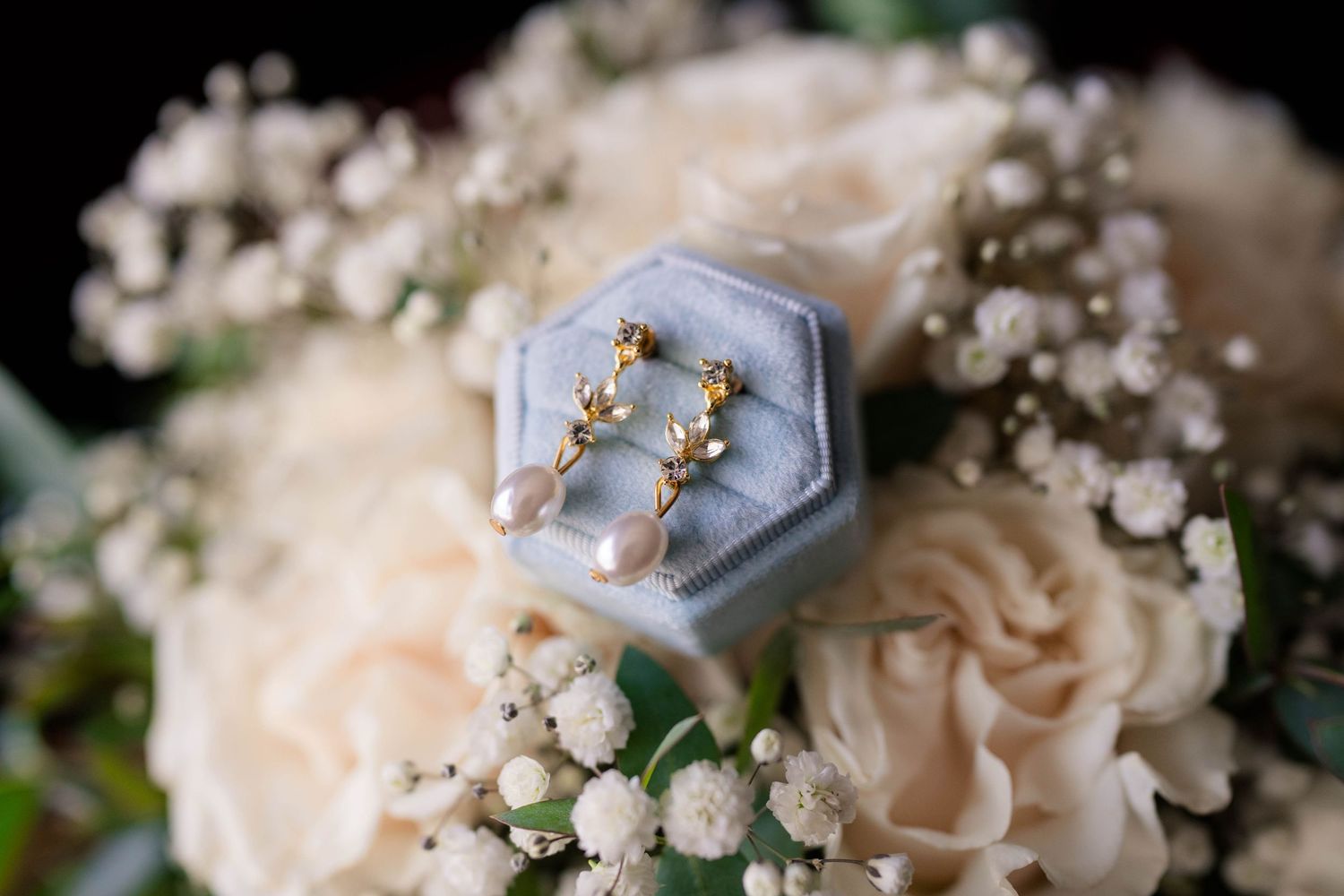 Gold drop earrings with pearl accents displayed in a blue velvet ring box surrounded by white roses and baby's breath.