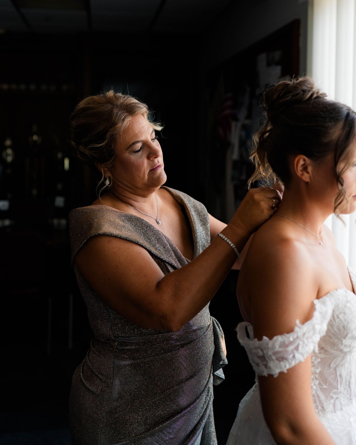 A woman in silver dress carefully fastens jewelry on a bride in an off-shoulder lace wedding gown.