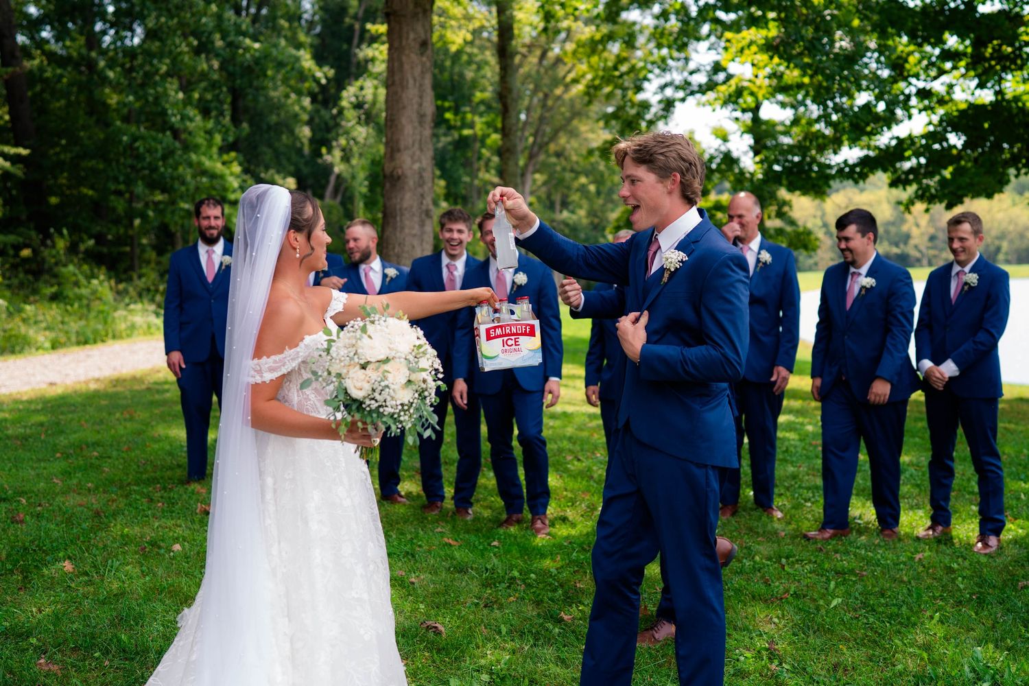 Bride and groom share a fun moment with wedding party dressed in navy suits and mauve bridesmaid dresses outdoors.
