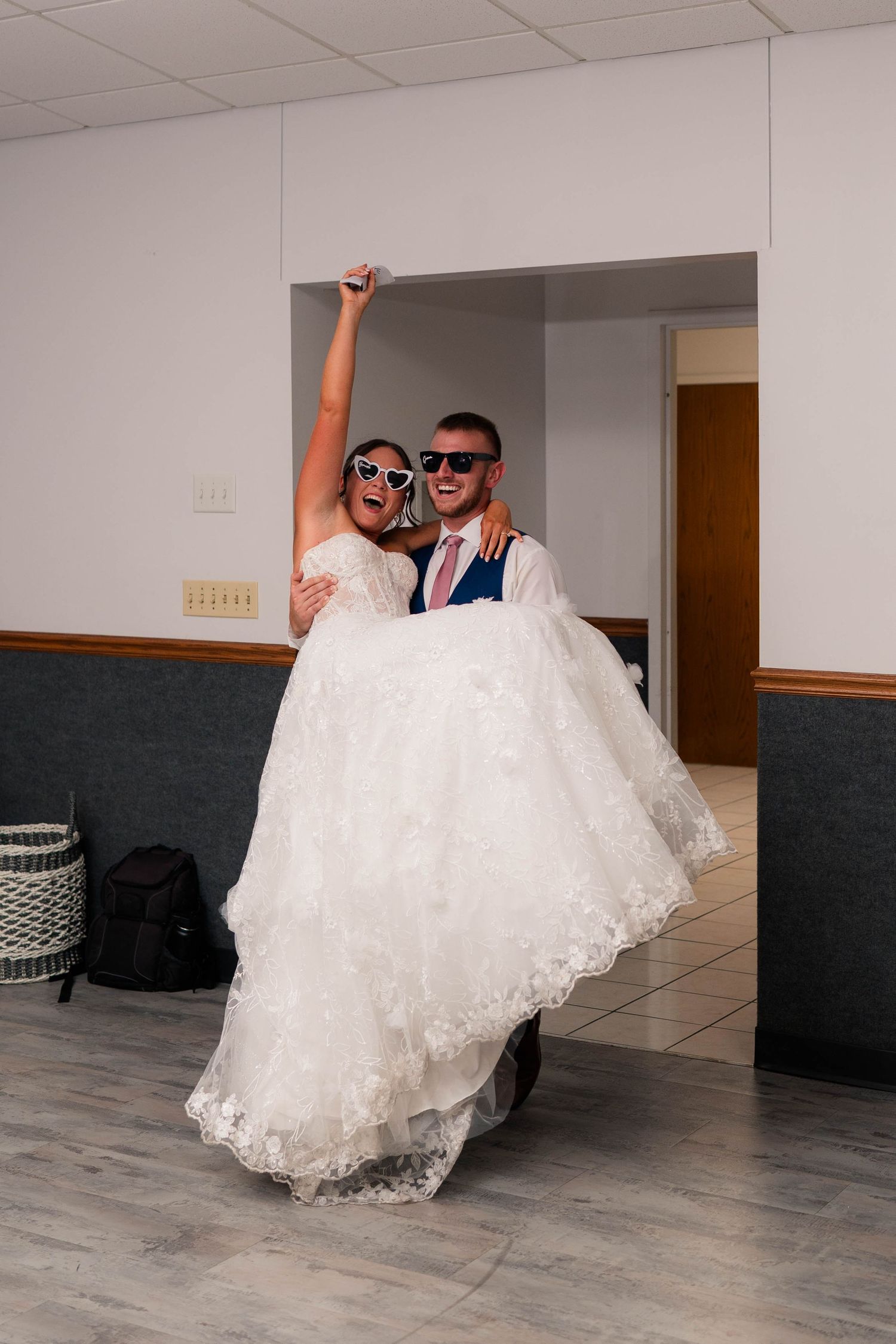 Groom carrying bride in white lace gown through doorway both wearing sunglasses celebrating after wedding.