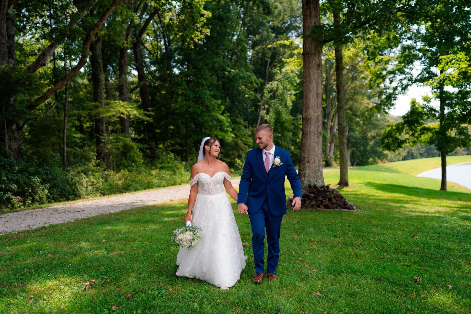 Bride and groom walking together on grassy path surrounded by tall trees on wedding day.