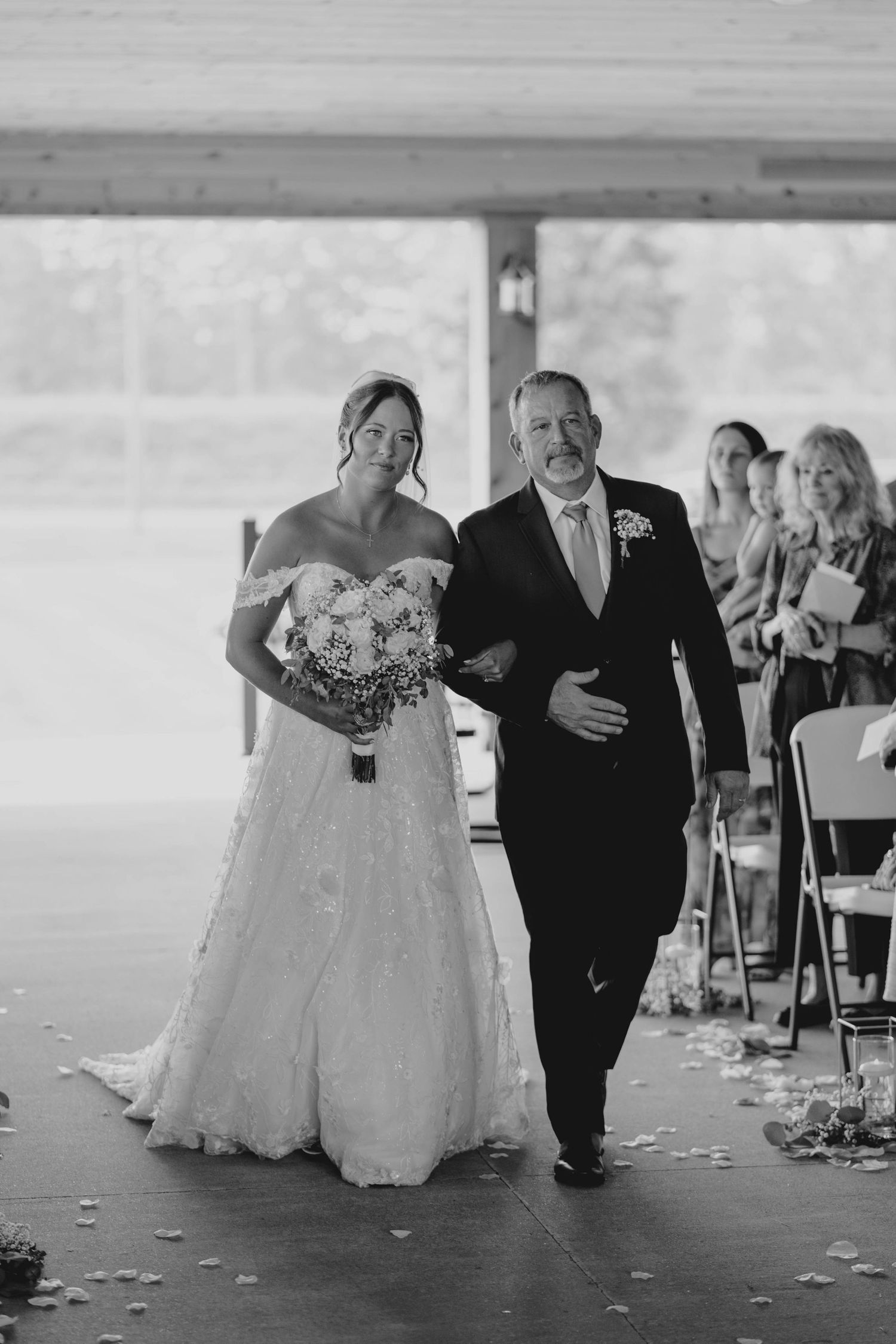 Black and white photo of a bride in a lace gown being walked down the aisle by her father.