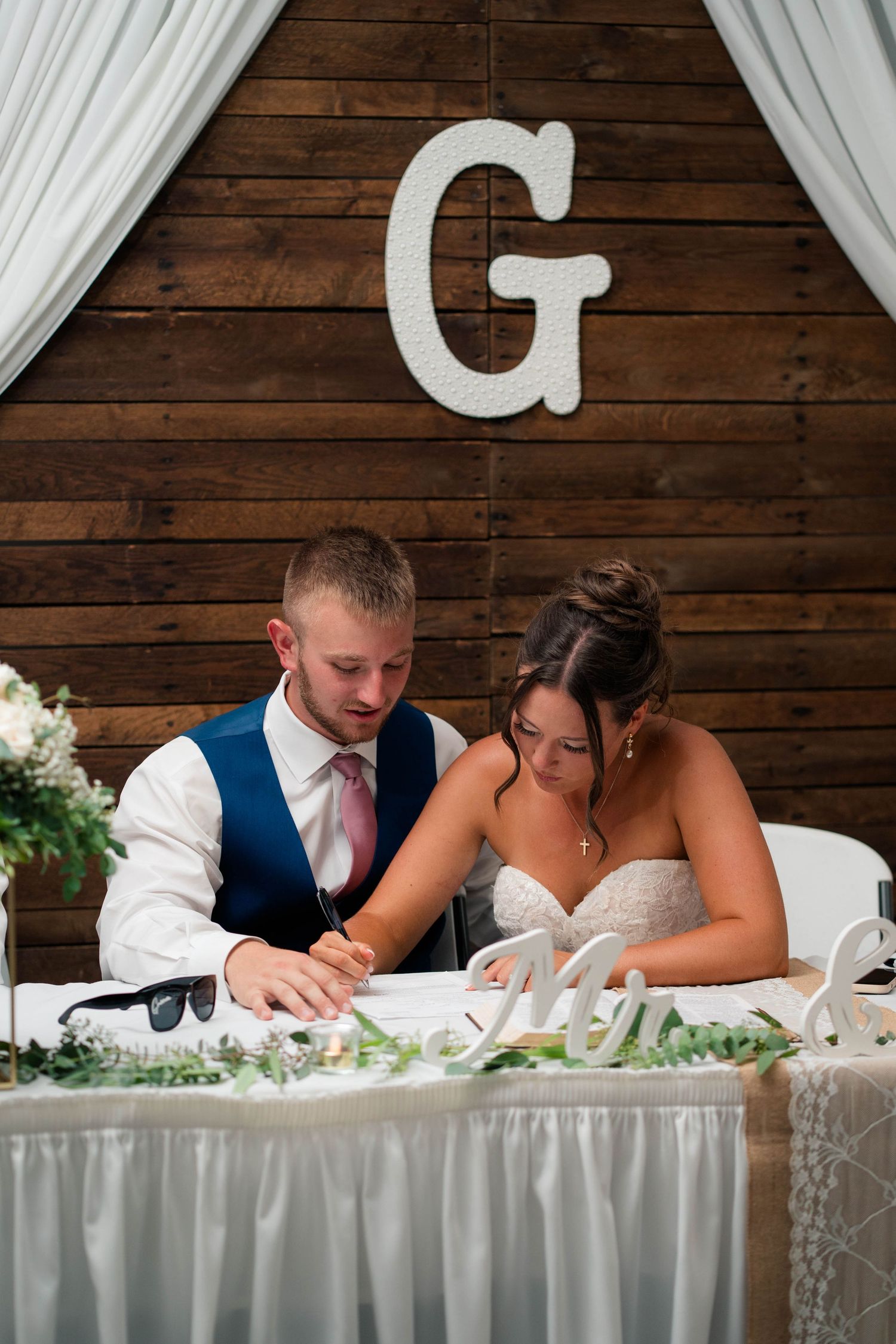 Bride and groom sign marriage certificate at sweetheart table with wooden G monogram backdrop.