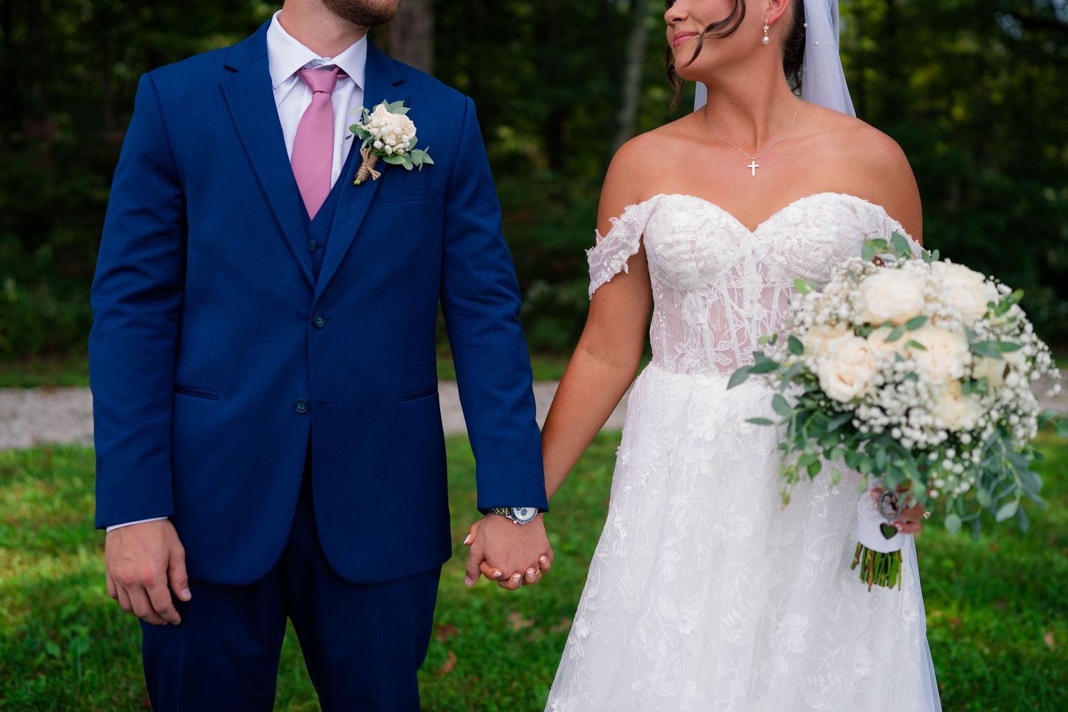 Bride in lace off-shoulder gown holds bouquet while groom in navy suit with pink tie holds her hand outdoors.