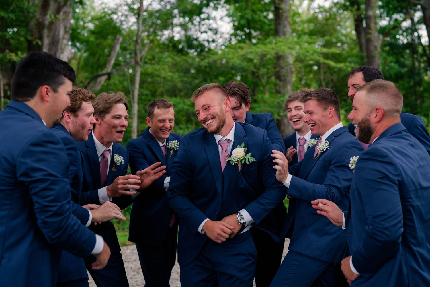 Groom laughs joyfully surrounded by groomsmen in matching navy suits during outdoor wedding celebration.